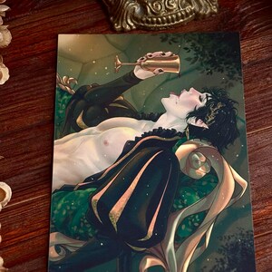 May include: An art print featuring a person reclining on a green and gold patterned chaise lounge, drinking from a gold goblet. The person is wearing a black outfit with gold accents. The background is a dark, stylized forest scene.