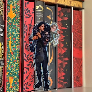 May include: A clear acrylic figure of a man carrying a woman, with a book collection in the background. The man is wearing a black outfit and boots. The woman has long hair and is wearing a green dress. The figure is on a clear stand.