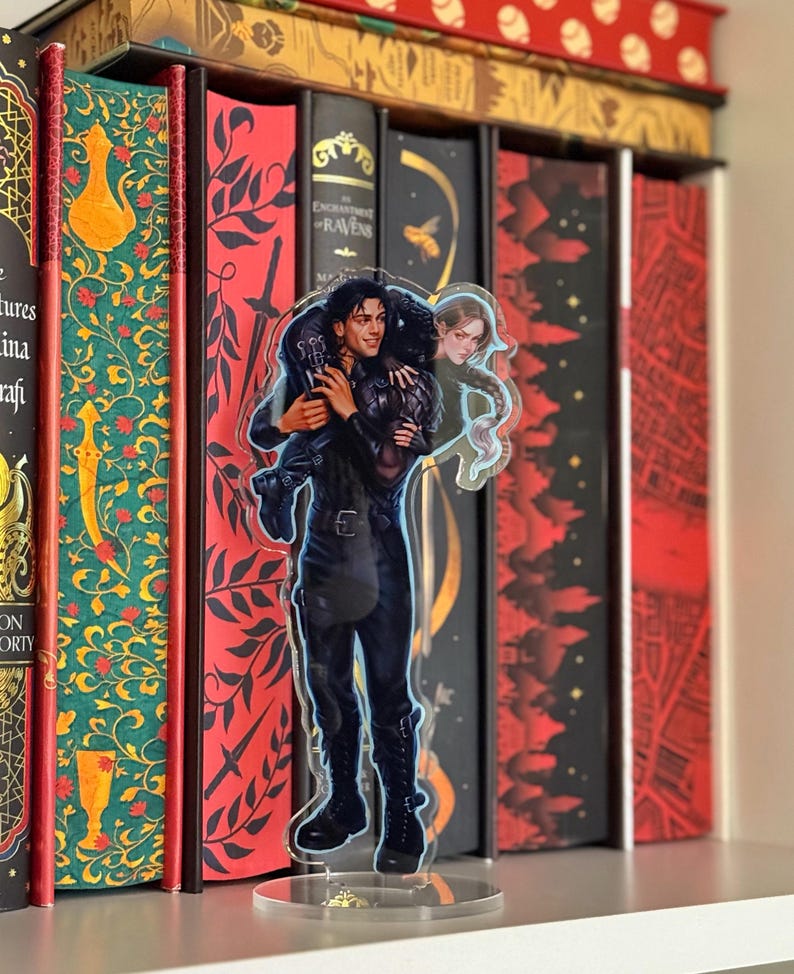 May include: A clear acrylic figure of a man carrying a woman, with a book collection in the background. The man is wearing a black outfit and boots. The woman has long hair and is wearing a green dress. The figure is on a clear stand.