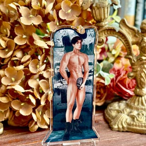 Puede incluir: Un soporte acrílico transparente con una figura masculina musculosa sosteniendo una calavera. La obra de arte tiene un fondo azul oscuro y gris. La figura lleva botas negras. El soporte está sobre una superficie de madera.