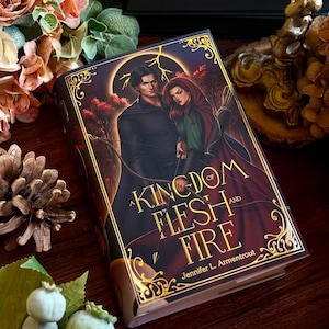May include: A hardback book titled "A Kingdom of Flesh and Fire" by Jennifer L. Armentrout. The cover depicts a couple, with gold decorative detailing. The book is surrounded by flowers and other decorative items, on a wooden surface.