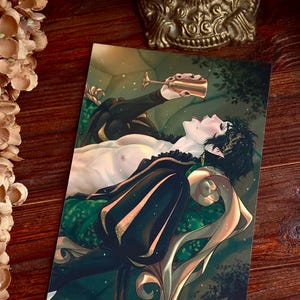 May include: Art print featuring a stylized illustration of a person reclining and drinking from a gold goblet. The person is wearing a black outfit with gold accents, and is lying on a green and gold patterned surface.