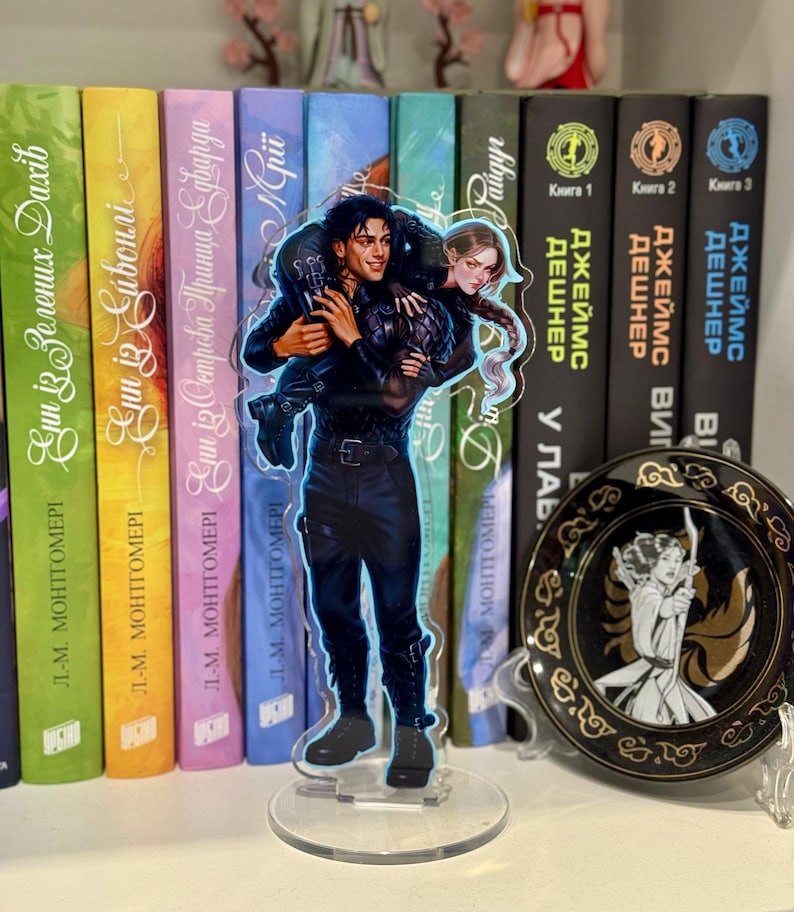 May include: A clear acrylic stand-up figure featuring a man carrying a woman. The man is dressed in black, and the woman has long hair. The figure is in front of a shelf of books with colorful spines. A decorative plate is also visible.