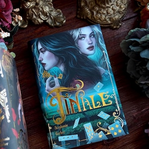 Puede incluir: Un libro titulado "Finale" de Stephanie Garber. La portada presenta a dos mujeres y naipes. El título es dorado. El fondo es un paisaje verde con cielo azul. El libro está sobre una superficie de madera oscura.