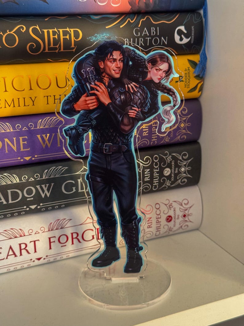May include: A clear acrylic stand-up featuring a detailed illustration of two characters. The male character is carrying the female character on his back. The artwork is set against a backdrop of colorful books.