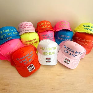 May include: A collection of ten colorful trucker hats with various phrases printed on them in neon colors. The phrases include "I'm fine it's just my RBF", "I shaved my bush for this", "I'd rather go home than go big", "Dibs on the redhead", "Hoochie daddy hunting", "I put the hot in psychotic", "I'm cool but I cry a lot", "Feral but friendly", "Sorry if that was weird. It'll probably happen again", and "Blink if you want me."