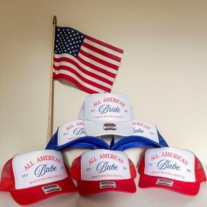 May include: A collection of trucker hats in red, white, and blue, with the text "ALL AMERICAN Babe" and "ALL AMERICAN Bride" in script. An American flag is displayed in the background, suggesting a patriotic theme.