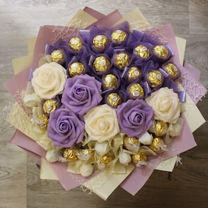 Puede incluir: Un ramo de flores artificiales con rosas moradas y crema, dispuestas alrededor de un grupo de caramelos de chocolate envueltos.