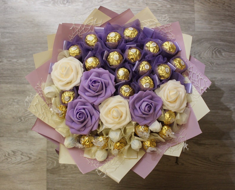 Puede incluir: Un ramo de flores artificiales con rosas moradas y crema, dispuestas alrededor de un grupo de caramelos de chocolate envueltos.