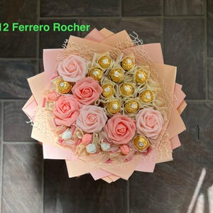 Puede incluir: Un ramo de rosas artificiales de color rosa con un centro de bombones Ferrero Rocher. El ramo est&aacute; envuelto en papel melocot&oacute;n con una red transparente superpuesta. El texto "12 Ferrero Rocher" es visible en la parte superior de la imagen.