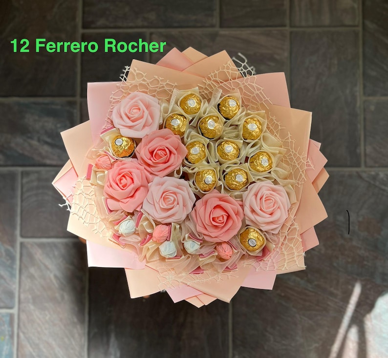 Puede incluir: Un ramo de rosas artificiales de color rosa con un centro de bombones Ferrero Rocher. El ramo est&aacute; envuelto en papel melocot&oacute;n con una red transparente superpuesta. El texto "12 Ferrero Rocher" es visible en la parte superior de la imagen.