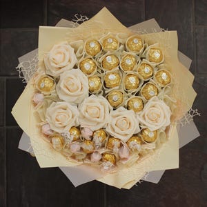 Puede incluir: Un ramo de rosas blancas y chocolates envueltos en oro dispuestos en un envoltorio de papel crema.