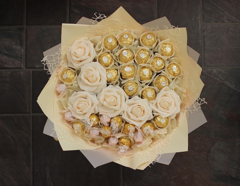 Puede incluir: Un ramo de rosas blancas y chocolates envueltos en oro dispuestos en un envoltorio de papel crema.