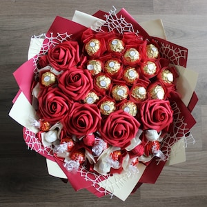 Puede incluir: Un ramo de rosas rojas y chocolates envueltos en papel de oro. El ramo est&aacute; atado con una cinta blanca y decorado con una puntilla blanca.