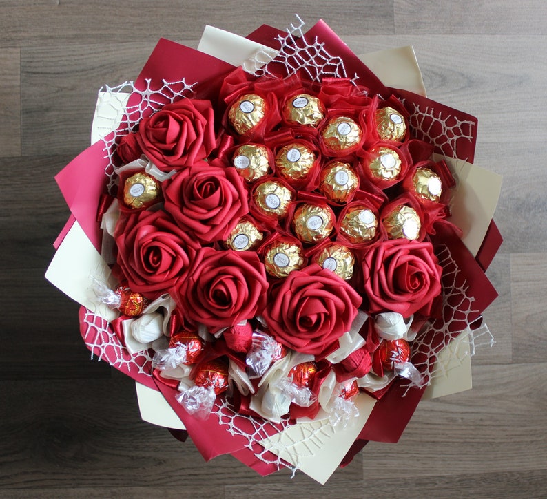 Puede incluir: Un ramo de rosas rojas y chocolates envueltos en papel de oro. El ramo est&aacute; atado con una cinta blanca y decorado con una puntilla blanca.