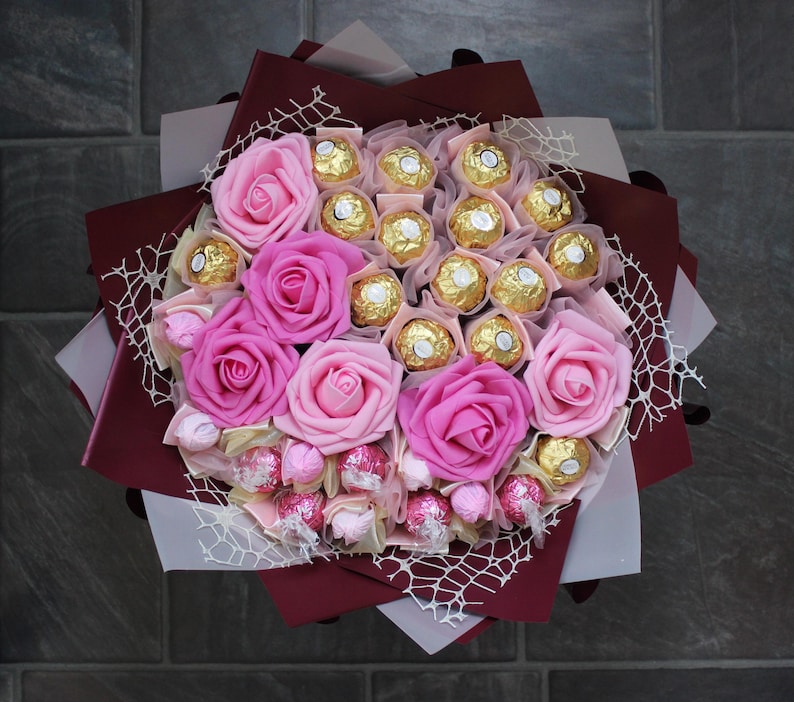 Puede incluir: Un ramo de rosas rosas y trufas de chocolate envueltas en papel burdeos y una red blanca.
