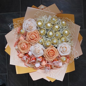 Puede incluir: Un ramo de rosas artificiales de color melocot&oacute;n y blanco con un centro de chocolates surtidos envueltos en papel de oro.
