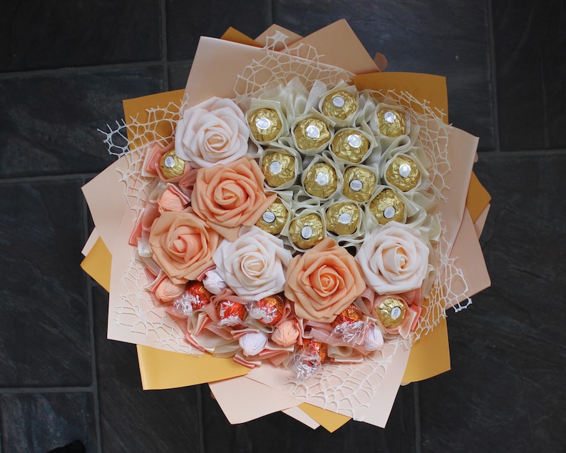 Puede incluir: Un ramo de rosas artificiales de color melocot&oacute;n y blanco con un centro de chocolates surtidos envueltos en papel de oro.