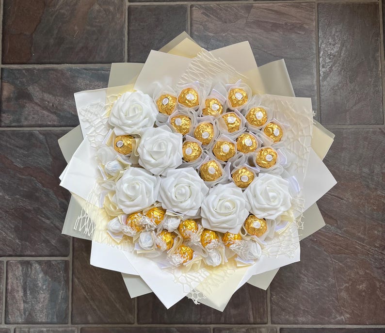 Puede incluir: Un ramo de rosas blancas de seda con un centro de chocolates envueltos en oro.