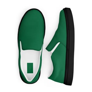 May include: A pair of green slip-on shoes with black soles and white interior lining. The shoes are shown from a top-down perspective, with one shoe slightly angled to the side. The shoes have a minimalist design.