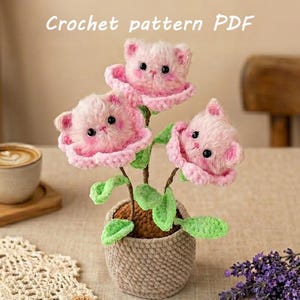 Op de afbeelding: Een gehaakte creatie met drie roze bloemen in de vorm van kittens in een geweven pot. De bloemen hebben groene bladeren. Bovenin staat de tekst "FLUFFY KITTEN FLOWERS Crochet pattern PDF".
