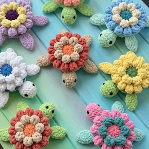 Può includere: Una collezione di tartarughe fatte a mano all'uncinetto, con gusci a forma di fiore di diversi colori. Le tartarughe hanno pinne e teste verdi e sono disposte su una superficie di legno blu e verde. I colori dei fiori includono viola, giallo, blu, bianco, arancione, rosso e rosa.