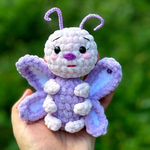 May include: A handmade, plush butterfly toy in shades of lavender, white, and pink. The butterfly has large, detailed wings, a round body, and delicate antennae. The toy is held in a hand against a blurred green background.