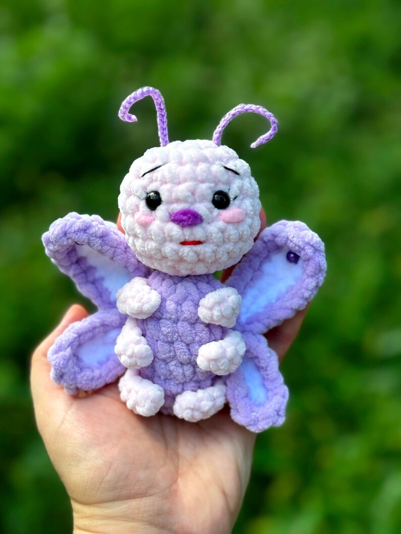 May include: A handmade, plush butterfly toy in shades of lavender, white, and pink. The butterfly has large, detailed wings, a round body, and delicate antennae. The toy is held in a hand against a blurred green background.