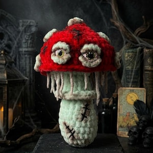 May include: A handcrafted crochet mushroom with a vibrant red cap and off-white spots. The mushroom features eerie, detailed eyes and a light green stem with stitched details, creating a spooky, whimsical aesthetic. It sits on a dark, textured surface.