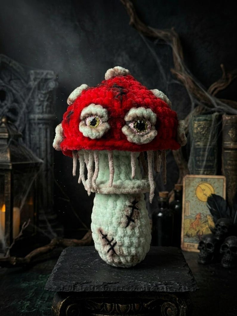 May include: A handcrafted crochet mushroom with a vibrant red cap and off-white spots. The mushroom features eerie, detailed eyes and a light green stem with stitched details, creating a spooky, whimsical aesthetic. It sits on a dark, textured surface.