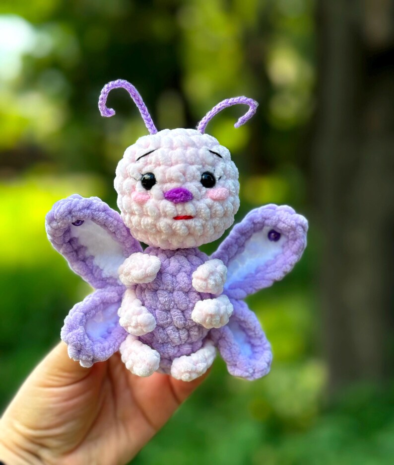 May include: A handmade crochet butterfly in shades of lavender, white, and pink. The plush toy features large wings, a round body, and delicate antennae. The butterfly has a sweet expression with black eyes and a small red mouth.