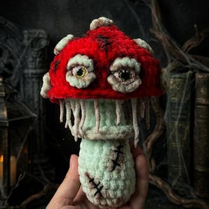 May include: A handmade, plush mushroom with a vibrant red cap and a light green stem. The cap features two large, detailed eyes and is trimmed with a lighter shade of green. The stem has stitched details, creating a spooky, Halloween-themed decoration.