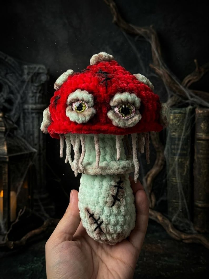 May include: A handmade, plush mushroom with a vibrant red cap and a light green stem. The cap features two large, detailed eyes and is trimmed with a lighter shade of green. The stem has stitched details, creating a spooky, Halloween-themed decoration.