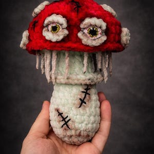 May include: A handmade, plush mushroom with a vibrant red cap and a light green stem. The mushroom features detailed, stitched eyes and decorative stitches, creating a unique, whimsical design. The mushroom is held in a hand.
