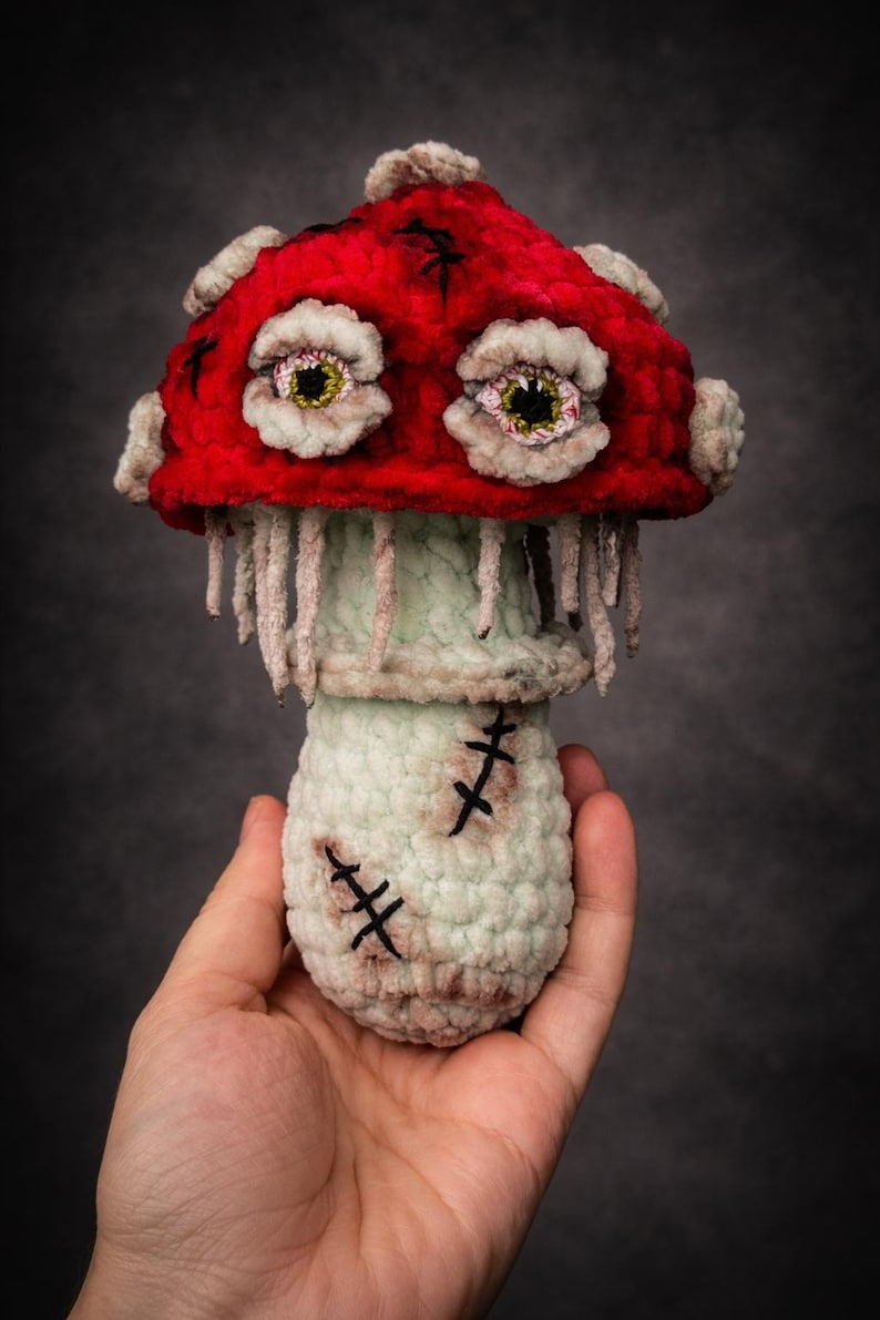 May include: A handmade, plush mushroom with a vibrant red cap and a light green stem. The mushroom features detailed, stitched eyes and decorative stitches, creating a unique, whimsical design. The mushroom is held in a hand.