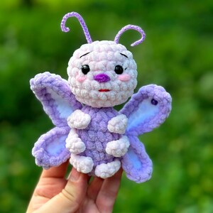 May include: A handmade crochet butterfly in shades of purple and white. The butterfly has large, detailed wings, a round body, and delicate antennae. The butterfly's face features small black eyes, pink cheeks, and a purple nose and mouth. The toy is held against a blurred green background.