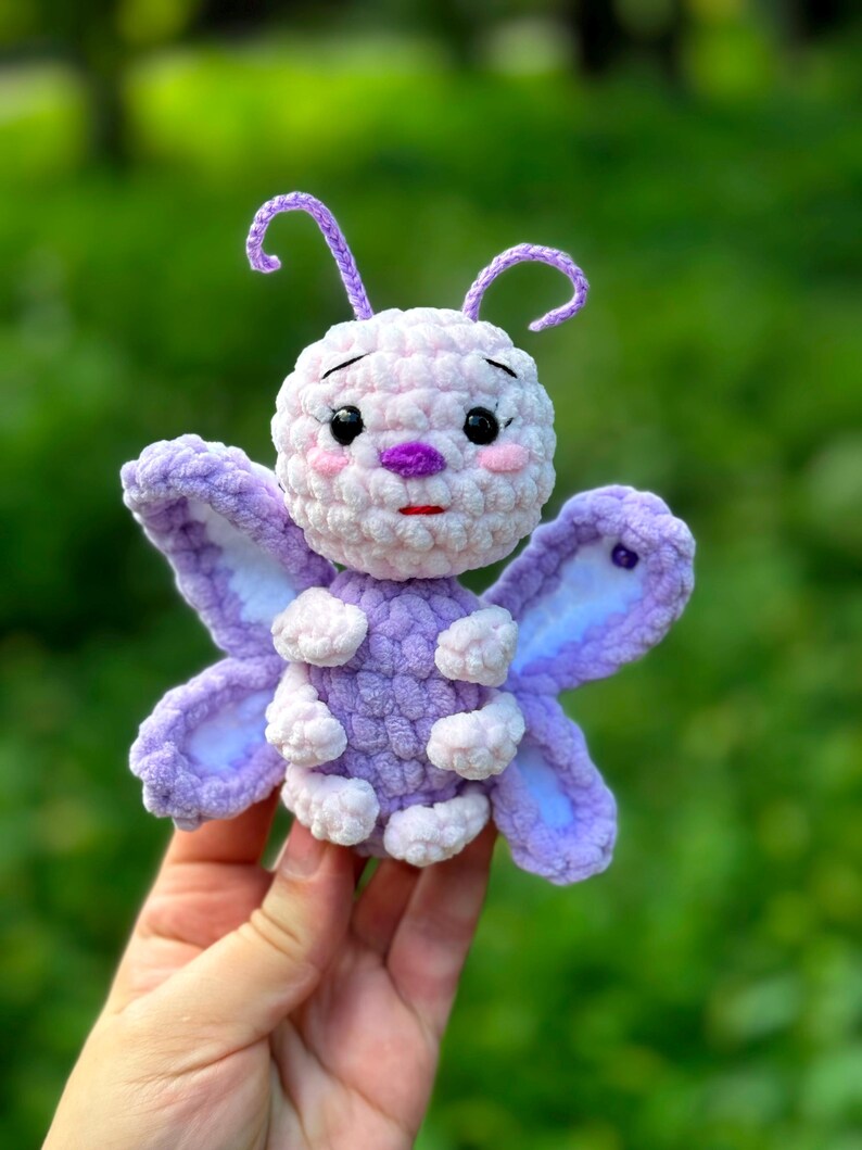 May include: A handmade crochet butterfly in shades of purple and white. The butterfly has large, detailed wings, a round body, and delicate antennae. The butterfly's face features small black eyes, pink cheeks, and a purple nose and mouth. The toy is held against a blurred green background.