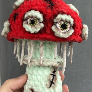 May include: A handmade, plush mushroom with a red cap, adorned with stitched black accents and two large, detailed eyes. The stem is light green with hanging strands, and the base features stitched details. A unique, whimsical decorative item.