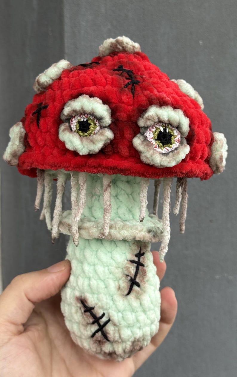 May include: A handmade, plush mushroom with a red cap, adorned with stitched black accents and two large, detailed eyes. The stem is light green with hanging strands, and the base features stitched details. A unique, whimsical decorative item.