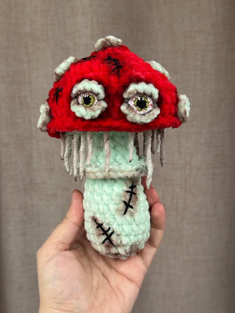 May include: A handmade, plush mushroom toy with a vibrant red cap and a light green stem and base. The mushroom features large, expressive eyes and stitched details, giving it a whimsical, slightly spooky appearance. The toy is held in a hand.