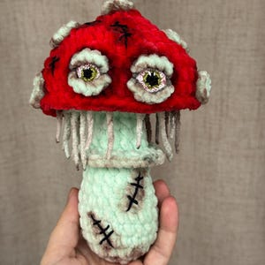 May include: A handmade plush mushroom with a red cap and mint green stem and base. The mushroom has two large, expressive eyes and is stitched with black thread. The mushroom is held in a hand.