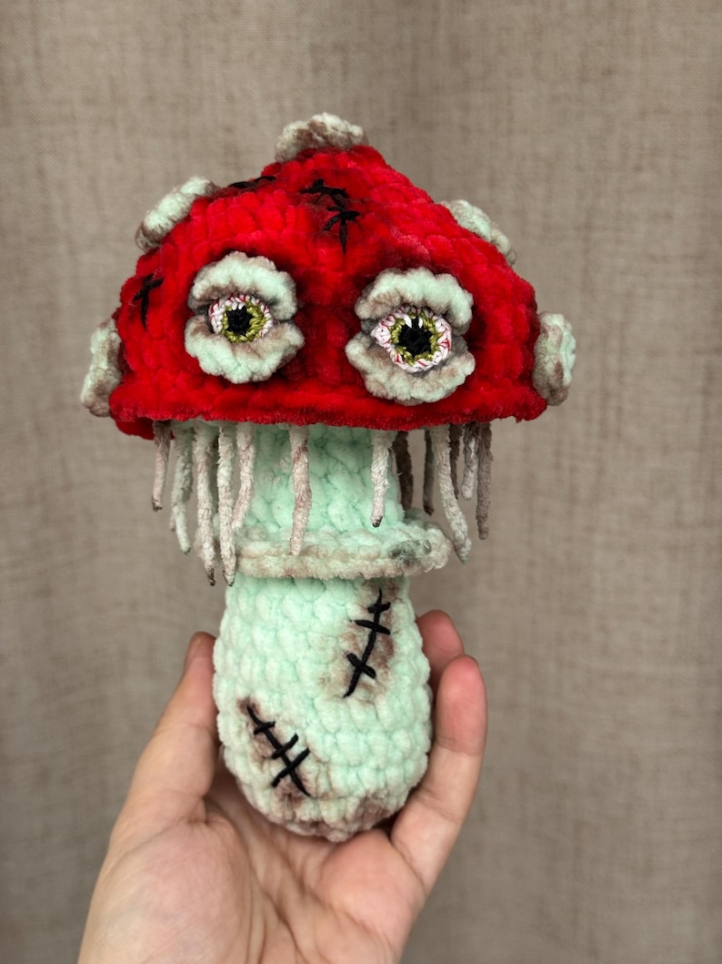 May include: A handmade plush mushroom with a red cap and mint green stem and base. The mushroom has two large, expressive eyes and is stitched with black thread. The mushroom is held in a hand.