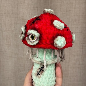 May include: A handmade crochet mushroom with a red cap and a light green stem. The cap features white spots and black stitches, with a stitched cross design. The mushroom has a spooky eye and dangling threads, creating a unique, whimsical design.