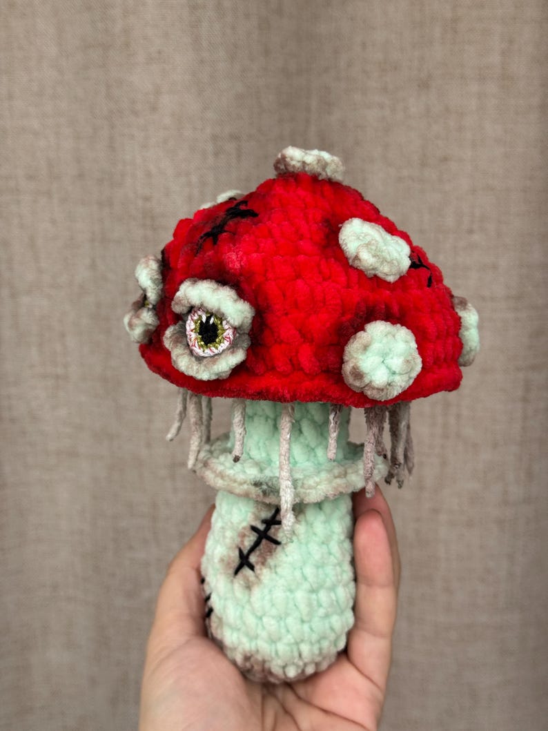 May include: A handmade crochet mushroom with a red cap and a light green stem. The cap features white spots and black stitches, with a stitched cross design. The mushroom has a spooky eye and dangling threads, creating a unique, whimsical design.