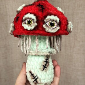May include: A handmade, plush mushroom figure with a red cap and a mint green stem. The cap features stitched details and two large, expressive eyes. The stem has stitched details and dangling threads, creating a unique, whimsical design.