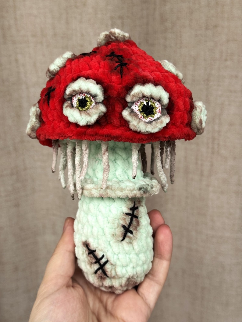 May include: A handmade, plush mushroom figure with a red cap and a mint green stem. The cap features stitched details and two large, expressive eyes. The stem has stitched details and dangling threads, creating a unique, whimsical design.