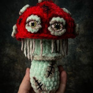 May include: A handmade crochet mushroom with a vibrant red cap and a light green stem. The mushroom features detailed, eerie eyes and stitched accents, creating a unique, whimsical design. The mushroom's stem has a stitched design.