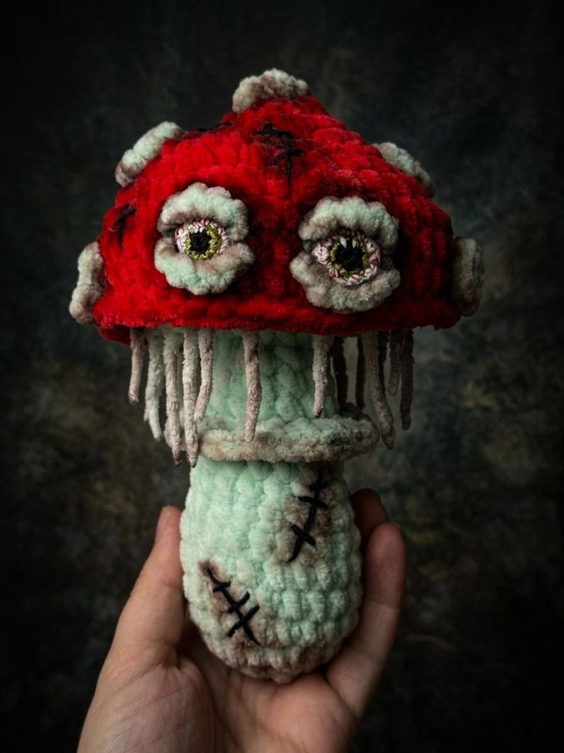 May include: A handmade crochet mushroom with a vibrant red cap and a light green stem. The mushroom features detailed, eerie eyes and stitched accents, creating a unique, whimsical design. The mushroom's stem has a stitched design.