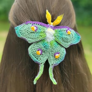 Könnte beinhalten: Ein handgefertigter Häkel-Haarclip in Form einer Mondmotte. Die Motte hat grüne, blaue und violette Flügel mit gelben und violetten Akzenten. Die Fühler sind gelb und der Körper ist weiß. Der Clip ist an braunem Haar befestigt.