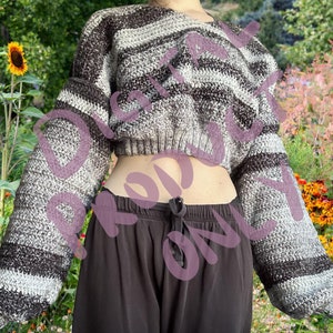 Pode incluir: Camisola de crochê cropped com mangas compridas. A camisola apresenta riscas horizontais em tons de cinza, castanho e branco. As mangas são largas e o corpo é curto. A camisola é feita à mão e tem um aspeto texturizado.